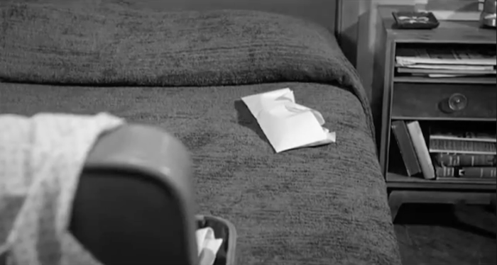 A slightly crumpled envelope on a bed, with a open suitcase in the lower left corner of the screen and a hotel bedside table on the right.