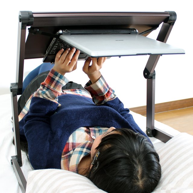 woman lying down on her back, reaching up and typing on a laptop held by a stand